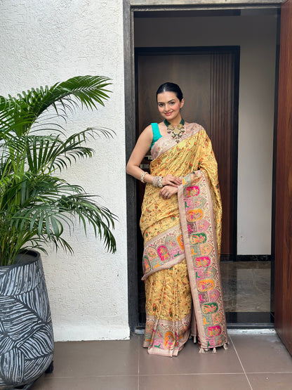 Woman in a traditional yellow saree with a colorful border standing indoors next to a plant. Peetambari Meenakari Jacquard Floral Print Saree | Yuvani Vesture