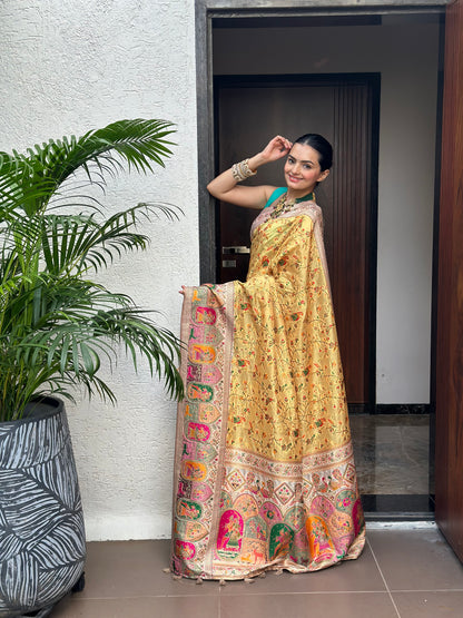 Woman in a gold saree with a colorful border standing indoors next to a plant. Peetambari Meenakari Jacquard Floral Print Saree | Yuvani Vesture